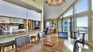 A spacious kitchen with white cabinets, a dining area with a large wooden table, and a view of the outdoors through large windows.