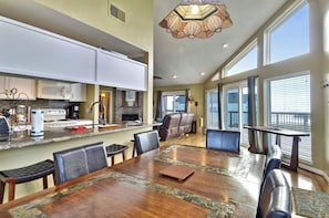 A spacious kitchen with white cabinets, a dining area with a large wooden table, and a view of the outdoors through large windows. - A Sunny Sandbar by Ryson Vacation Rentals (Galveston)