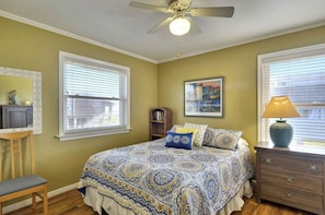 A bedroom with a large bed, a nightstand, a chair, a desk, a ceiling fan, and a window with blinds. - A Sunny Sandbar by Ryson Vacation Rentals (Galveston)