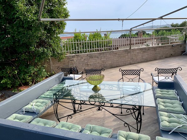A glass-top table with metal chairs and cushions, surrounded by a white fence, overlooking a body of water and greenery. - Letter Luxury Apartment (Formia)