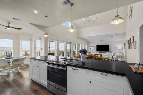 A spacious kitchen with a large island, white cabinetry, and a black countertop. There is a dining area with white chairs and a table, and a living room with a TV and seating.