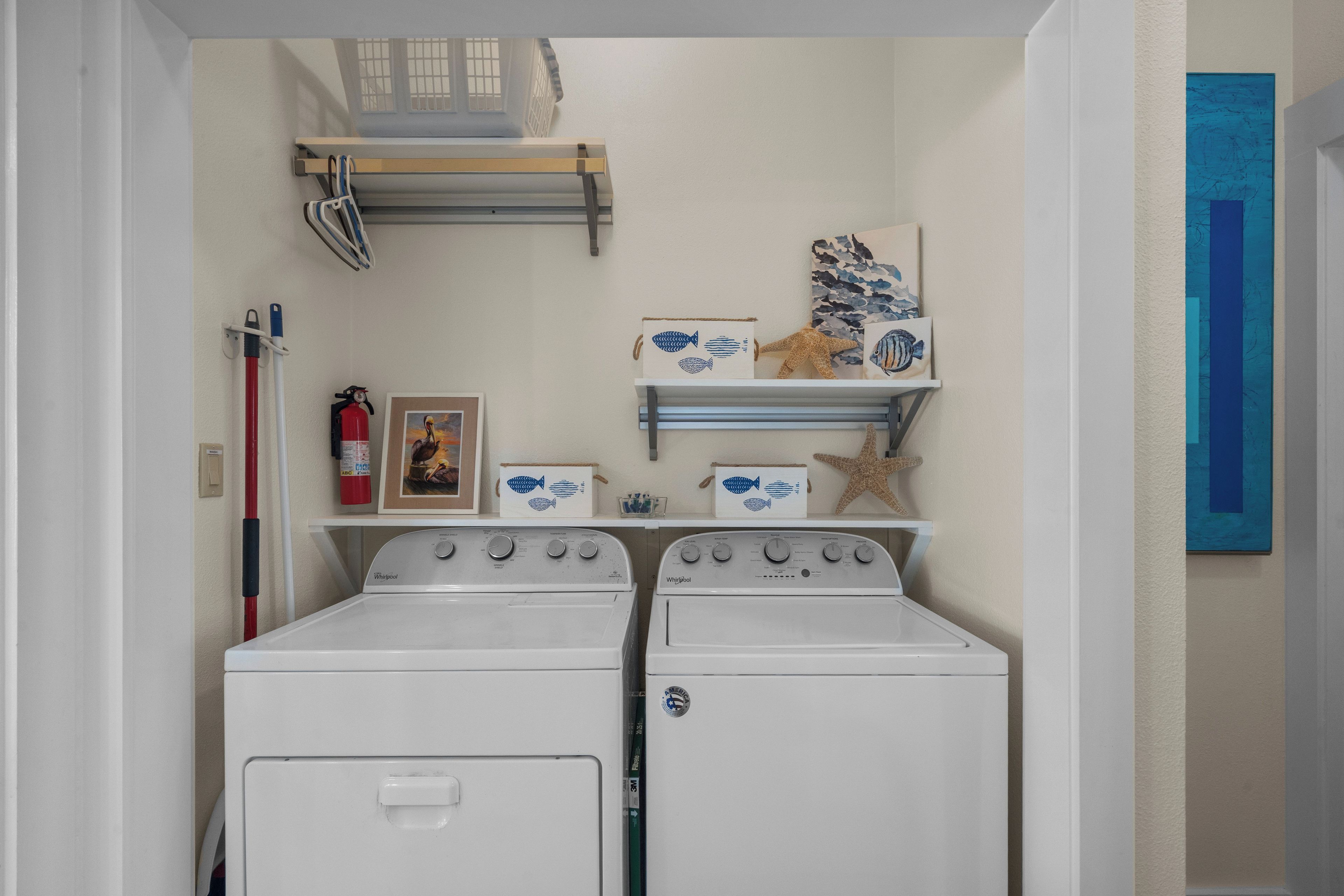 A laundry room with two white front-loading washers and dryers, a fire extinguisher, a framed picture, and decorative items on shelves.