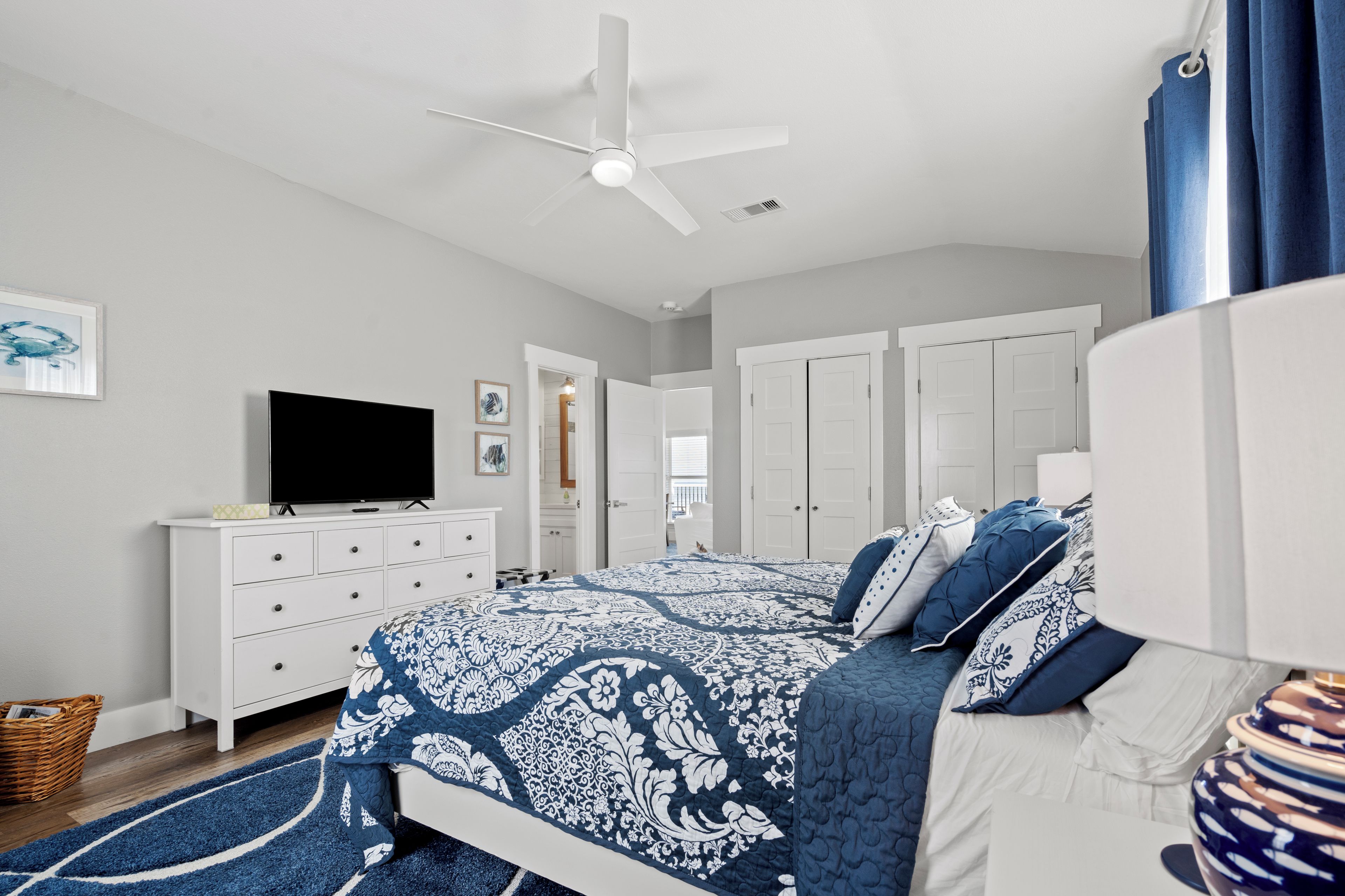 Une chambre à coucher avec un lit, un téléviseur posé sur une commode, un ventilateur de plafond et une porte donnant sur une autre pièce.
