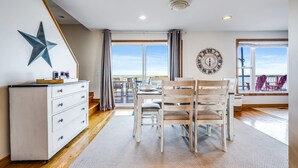Una habitación con piso de madera, un aparador blanco, una decoración con estrellas, un reloj de pared redondo y vista al mar a través de grandes ventanales.