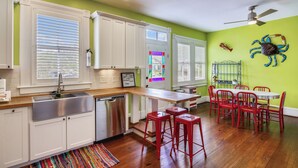 A kitchen with white cabinets, a stainless steel sink, and a wooden countertop. There is a red bar stool and a dining table with red chairs. - Abfab Seaside Cottage by Ryson Vacation Rentals (Galveston)