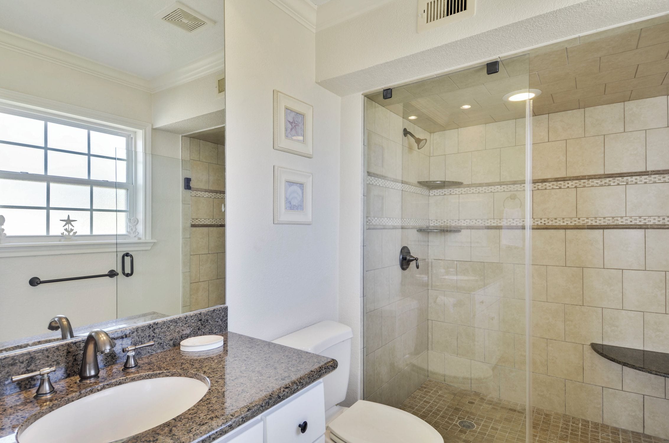 Une salle de bain avec un lavabo sur un plan de toilette en granit, des toilettes et une douche vitrée aux carreaux beiges.