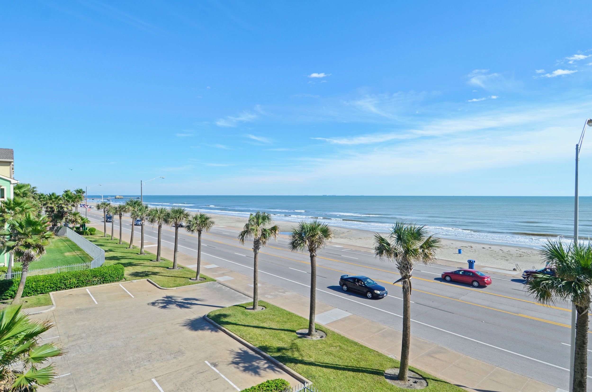 Una carretera costera con palmeras, un estacionamiento y una playa al fondo.