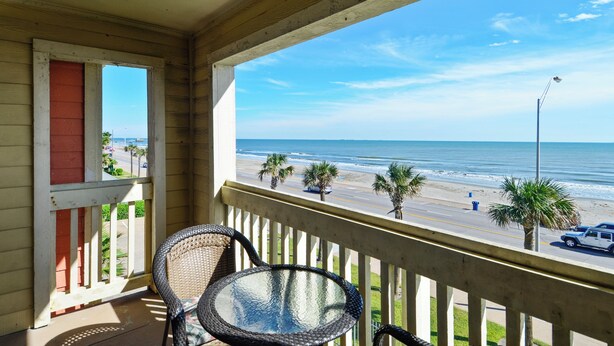 A balcony with wicker chairs and a glass table overlooking a beach with palm trees and a road. - Absolute Paradise-Dawn 1031 by Ryson Vacation Rentals (Galveston)