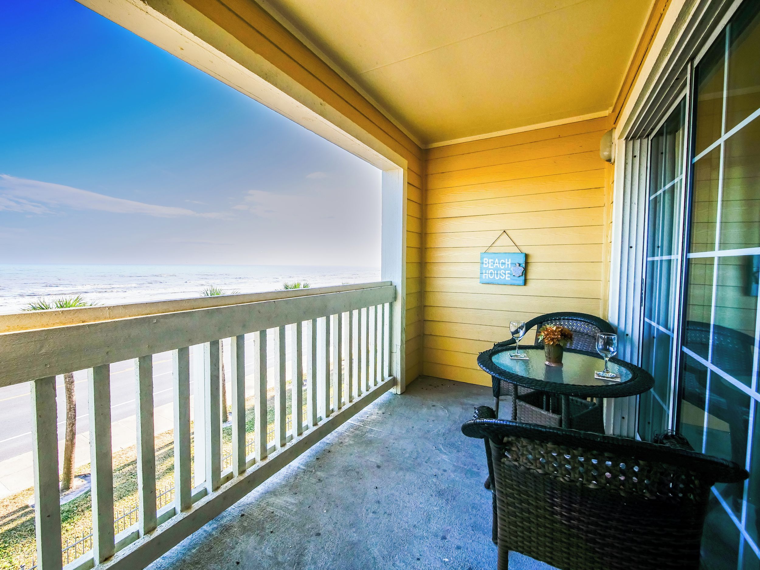 Uma varanda com vista para a praia, uma mesa com cadeiras e um letreiro 'Beach House' na parede.