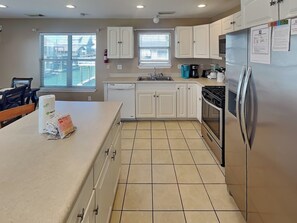 A kitchen with white cabinets, a stainless steel refrigerator, a dishwasher, and a window with blinds. - American Pride by Ryson Vacation Rentals (Galveston)