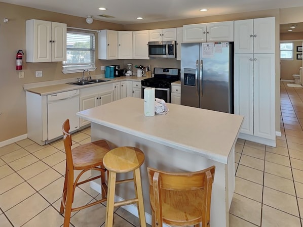 A kitchen with white cabinets, a central island with stools, a microwave, and a refrigerator. - American Pride by Ryson Vacation Rentals (Galveston)