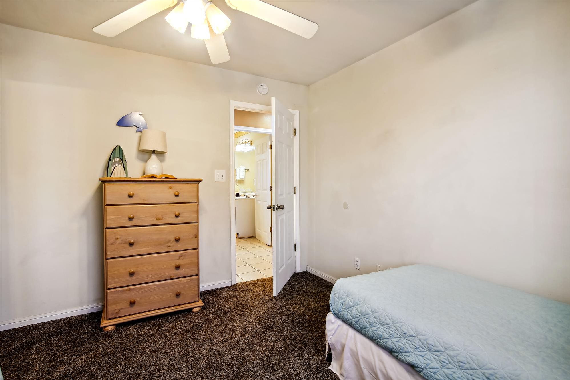 A bedroom with a wooden dresser, a bed with a blue quilt, and a ceiling fan.