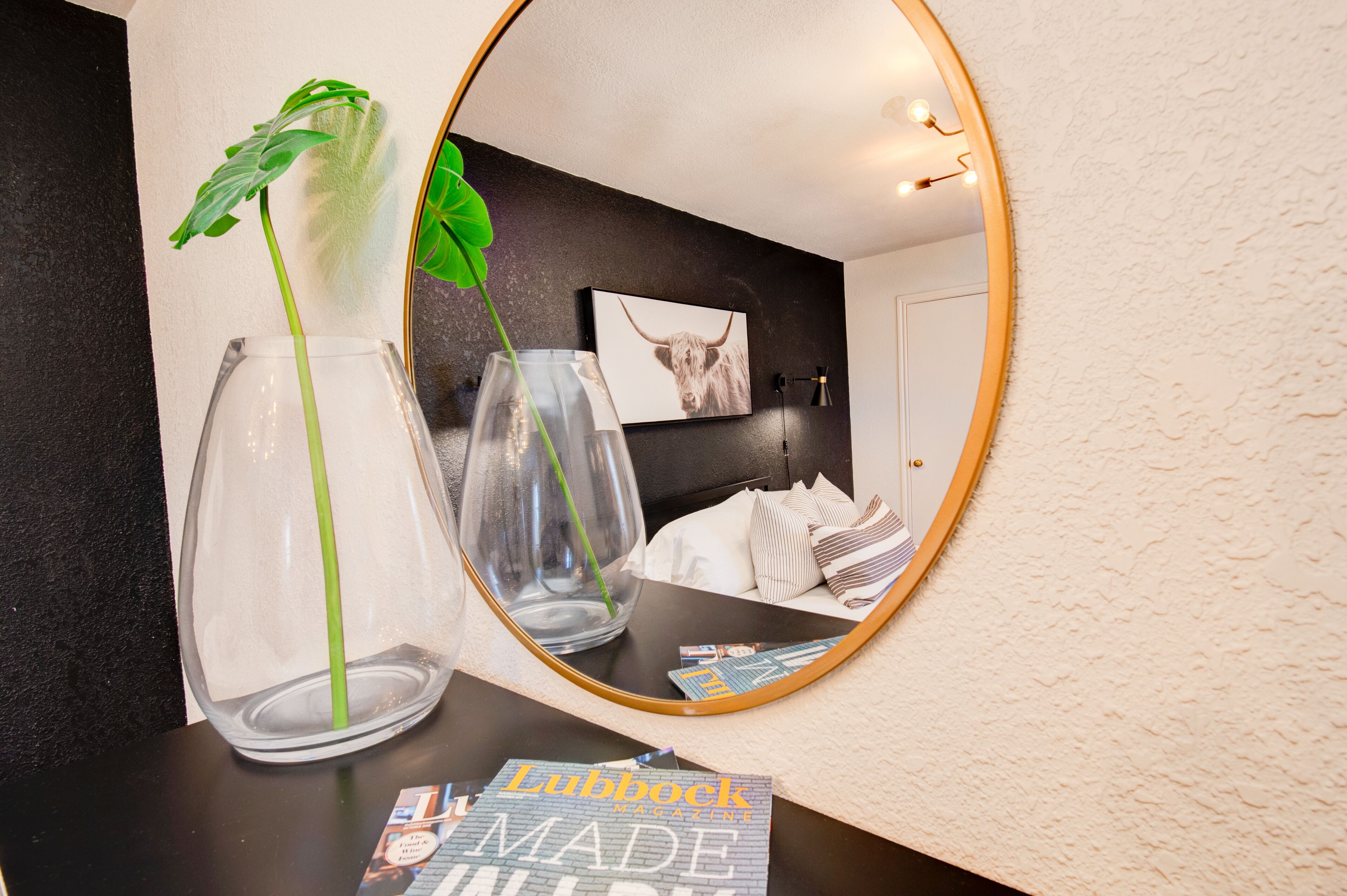 A room with a large mirror reflecting a bed, a framed picture of a bull, and a plant in a vase.