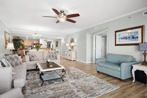 Ein Wohnzimmer mit Deckenventilator, Couchtisch, Sofa, einem hellblauen Sessel und einem Essbereich im Hintergrund.