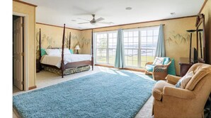 Een slaapkamer met een hemelbed, een groot raam met gordijnen, een plafondventilator en uitzicht op de omgeving.