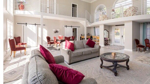 Een ruime woonkamer met een hoge plafond, een balkon en een eethoek met rode stoelen.