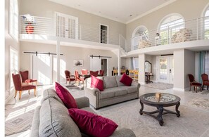 A spacious living area with a high ceiling, a balcony, and a dining area with red chairs.