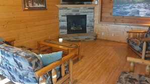 L’intérieur chaleureux d’un chalet avec une cheminée en pierre, des murs en bois, un téléviseur fixé au mur et un grand tableau représentant une scène de forêt.