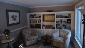 Un salon avec des fauteuils à rayures, une petite table ronde et une bibliothèque remplie de photos encadrées et d’objets décoratifs.