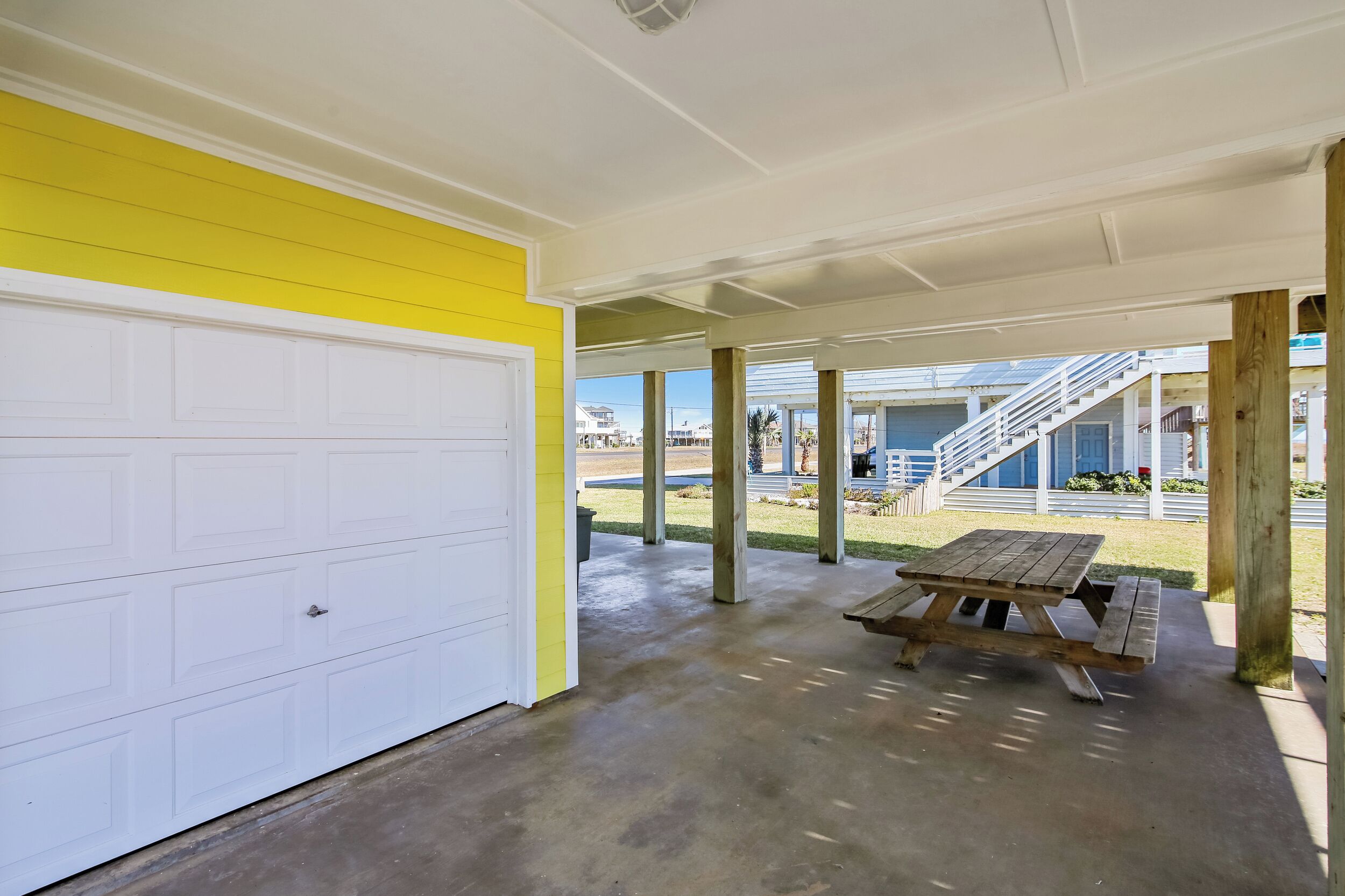 A covered outdoor area with a concrete floor, a wooden picnic table, and a white garage door.