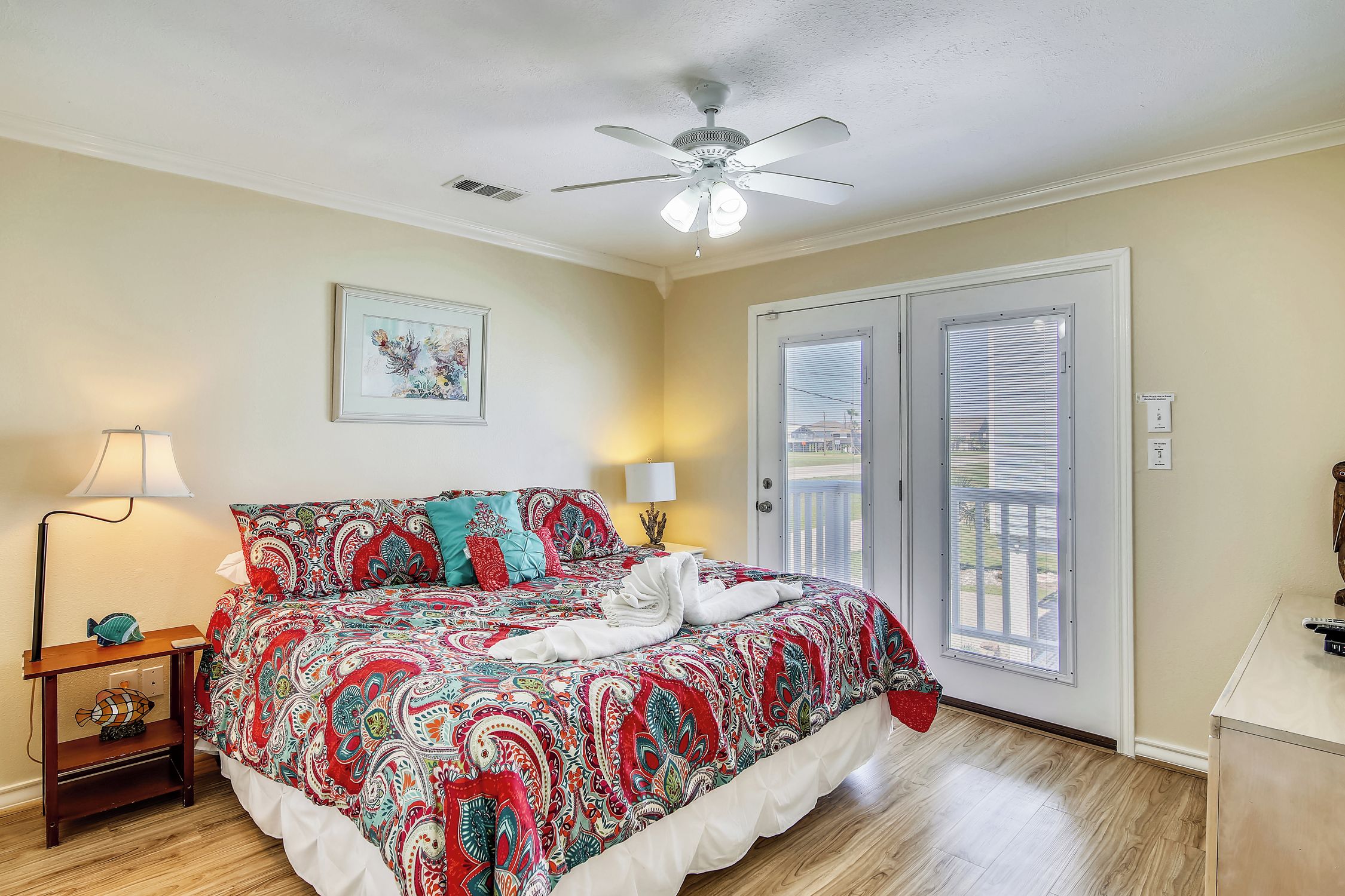 A bedroom with a bed, a ceiling fan, a nightstand with a lamp, and a door leading to a balcony.