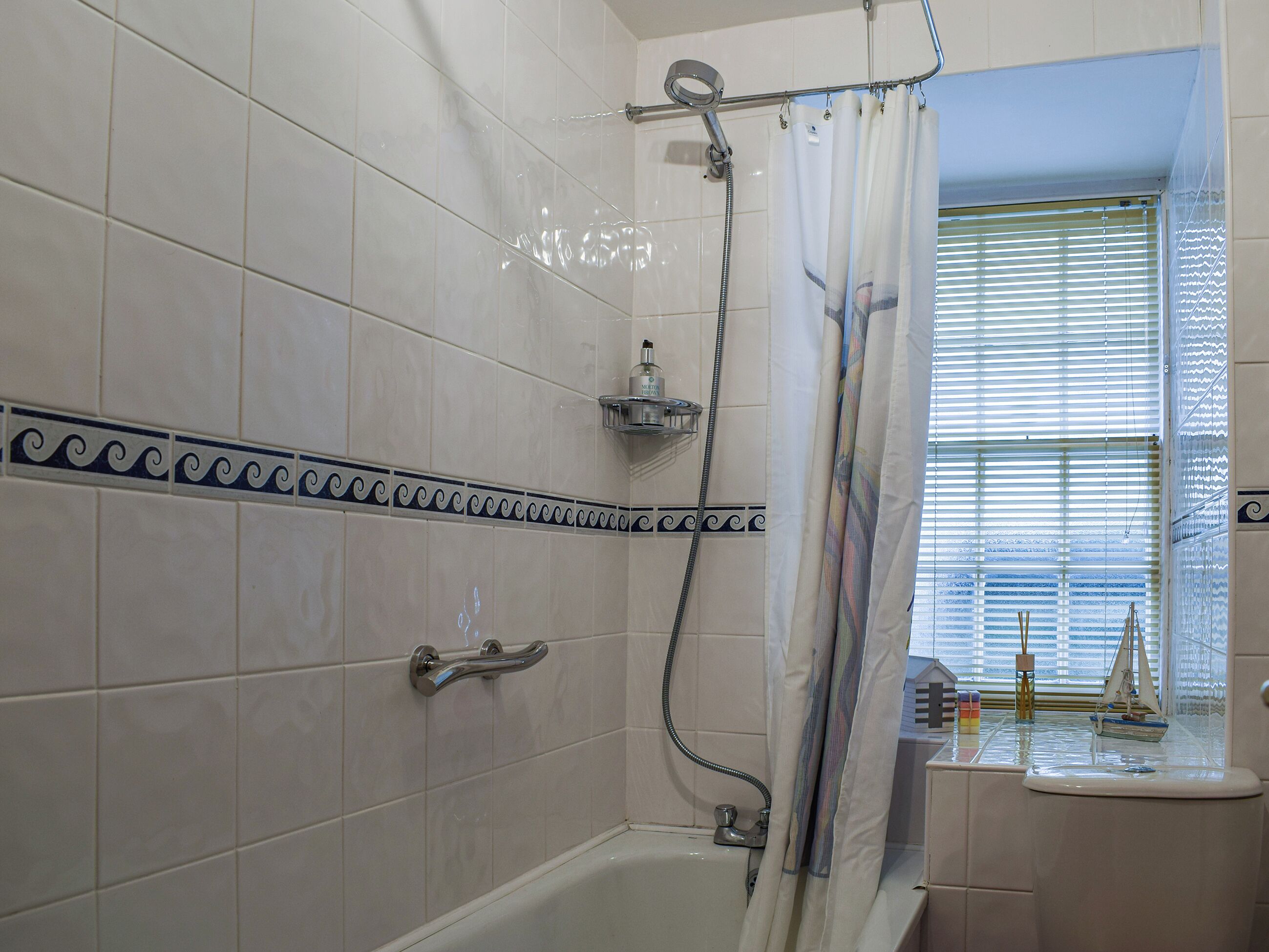 Ein Badezimmer mit weißen Fliesen, einer Duschkabine mit Vorhang, einer Badewanne, einem Fenster mit Jalousien und einer kleinen Ablage für Toilettenartikel.