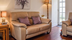 Ein Wohnzimmer mit einem beigefarbenen Sofa, zwei violetten Kissen, einer Stehlampe, einem Gemälde und einem kleinen Tisch mit Lampe und Waage.