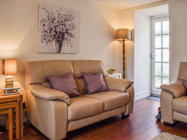 A living room with a beige sofa, two purple cushions, a floor lamp, a painting, and a small table with a lamp and a scale.