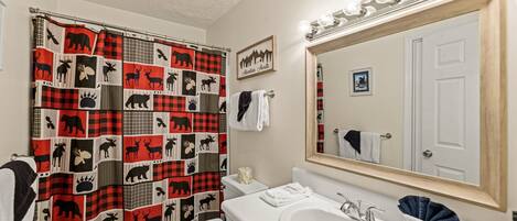 A bathroom with a red and black checkered shower curtain, a white sink, a mirror with lights, and a framed picture on the wall.