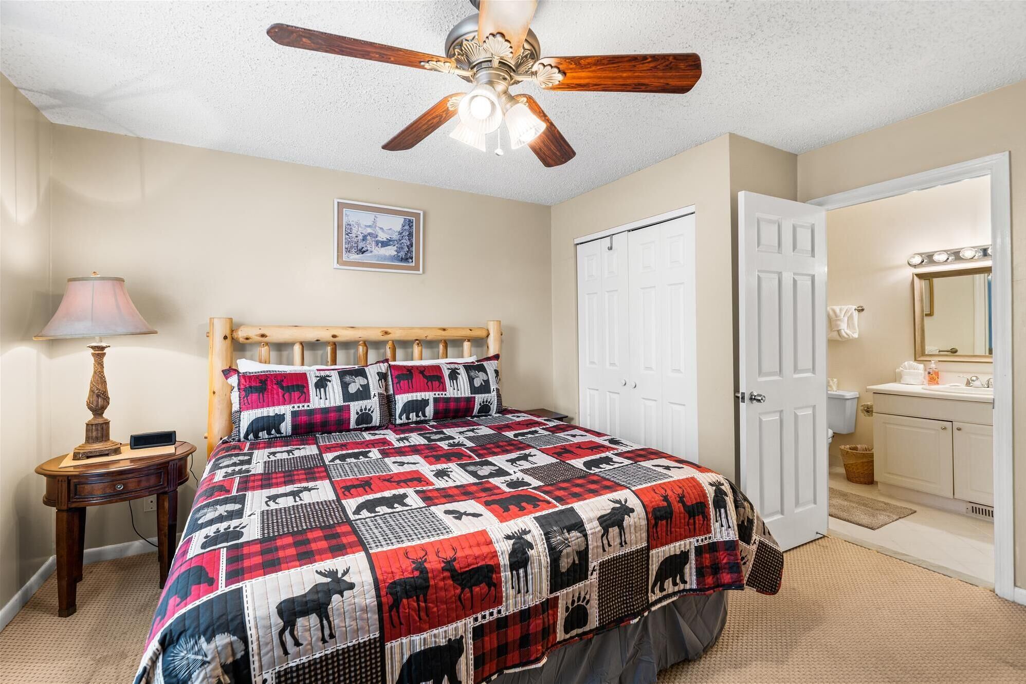 A bedroom with a wooden bed, a ceiling fan, a bedside table with a lamp, a quilt with animal prints, a door, and a bathroom visible through an open door.