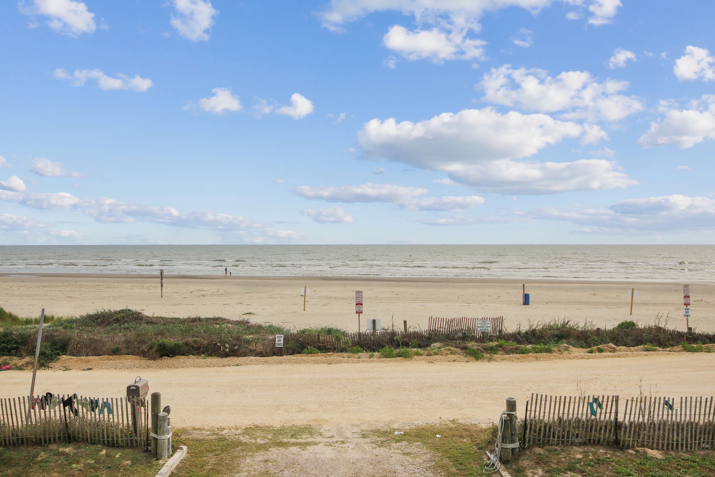 A beach with a sandy path leading to the sea, wooden fences, and a clear sky.