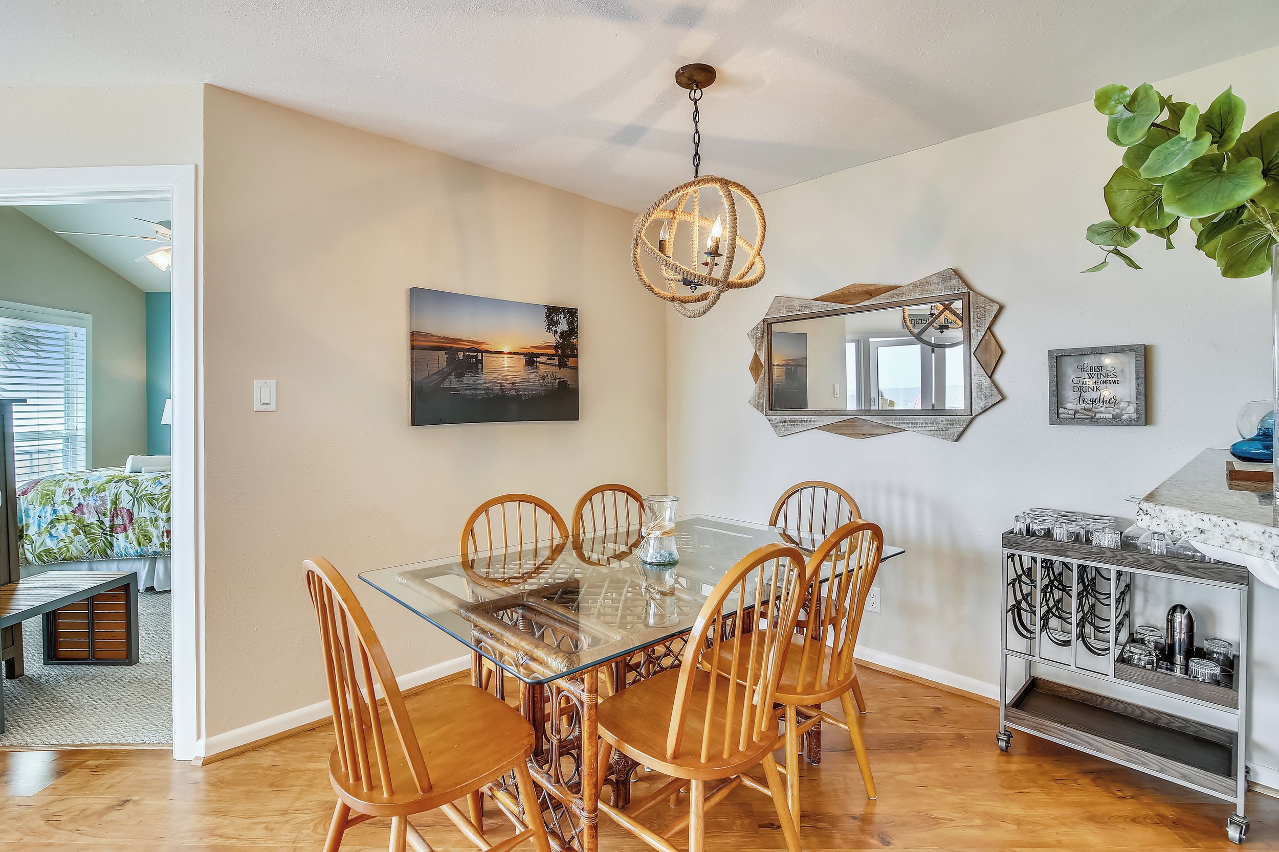 A dining area with a glass table, wooden chairs, a hanging light fixture, a wall-mounted mirror, and a cabinet with glassware.