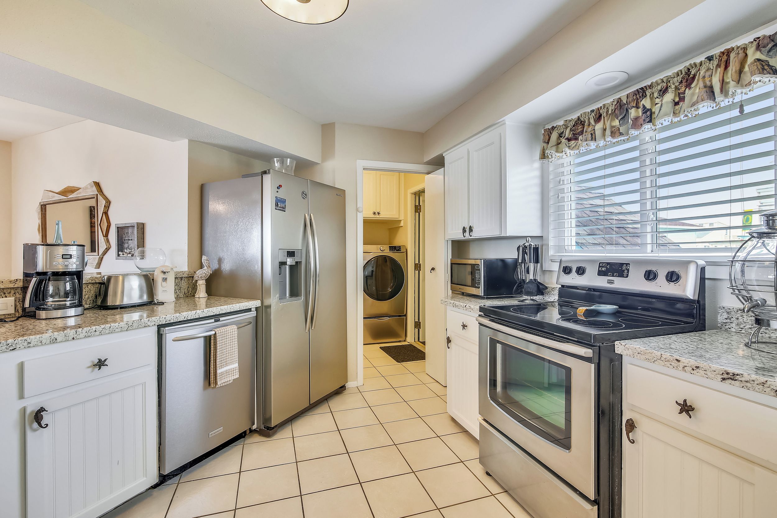A modern kitchen with stainless steel appliances, white cabinetry, and a large window with blinds.