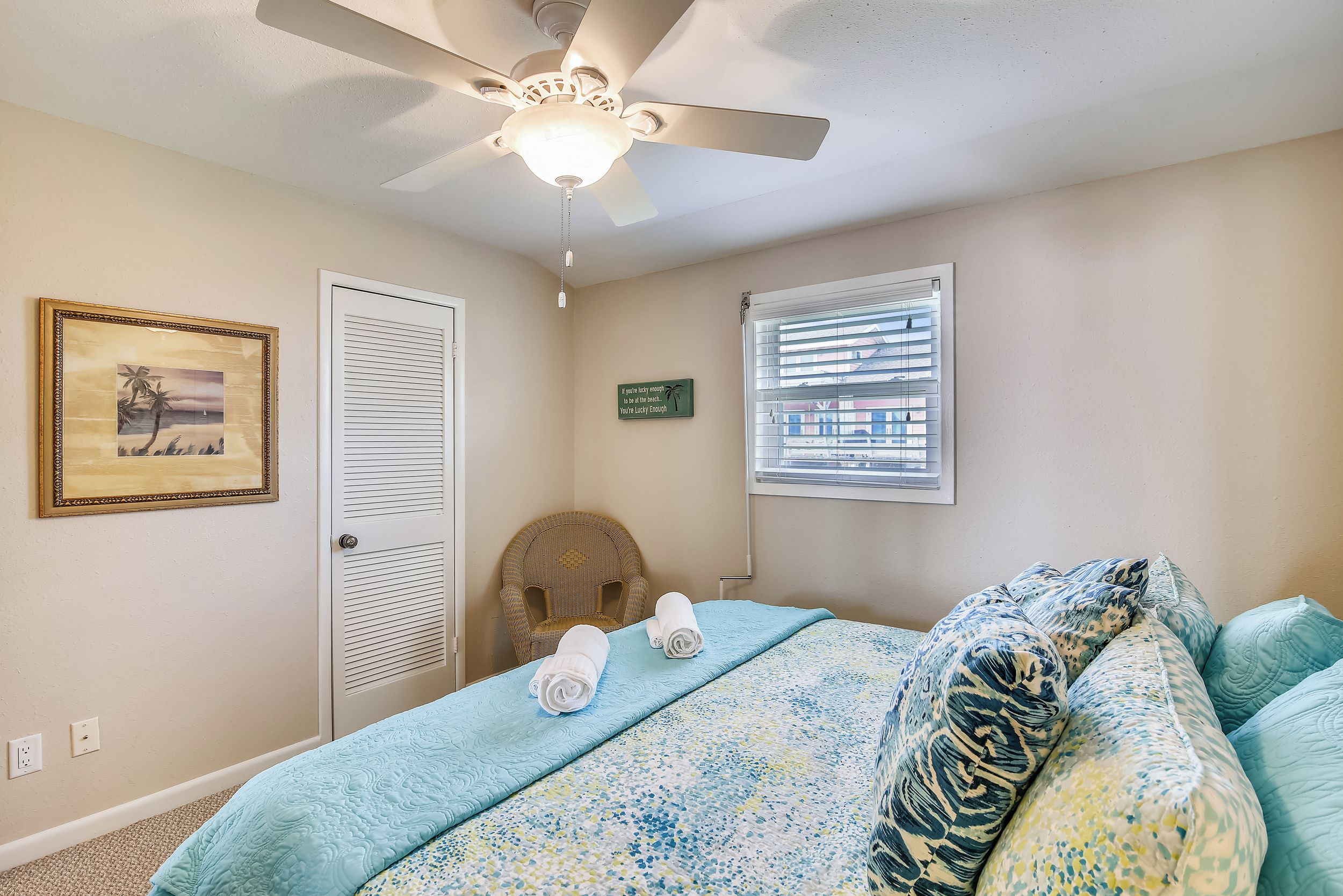 A bedroom with a bed, a ceiling fan, a door, a window with blinds, a framed picture, and a sign on the wall.