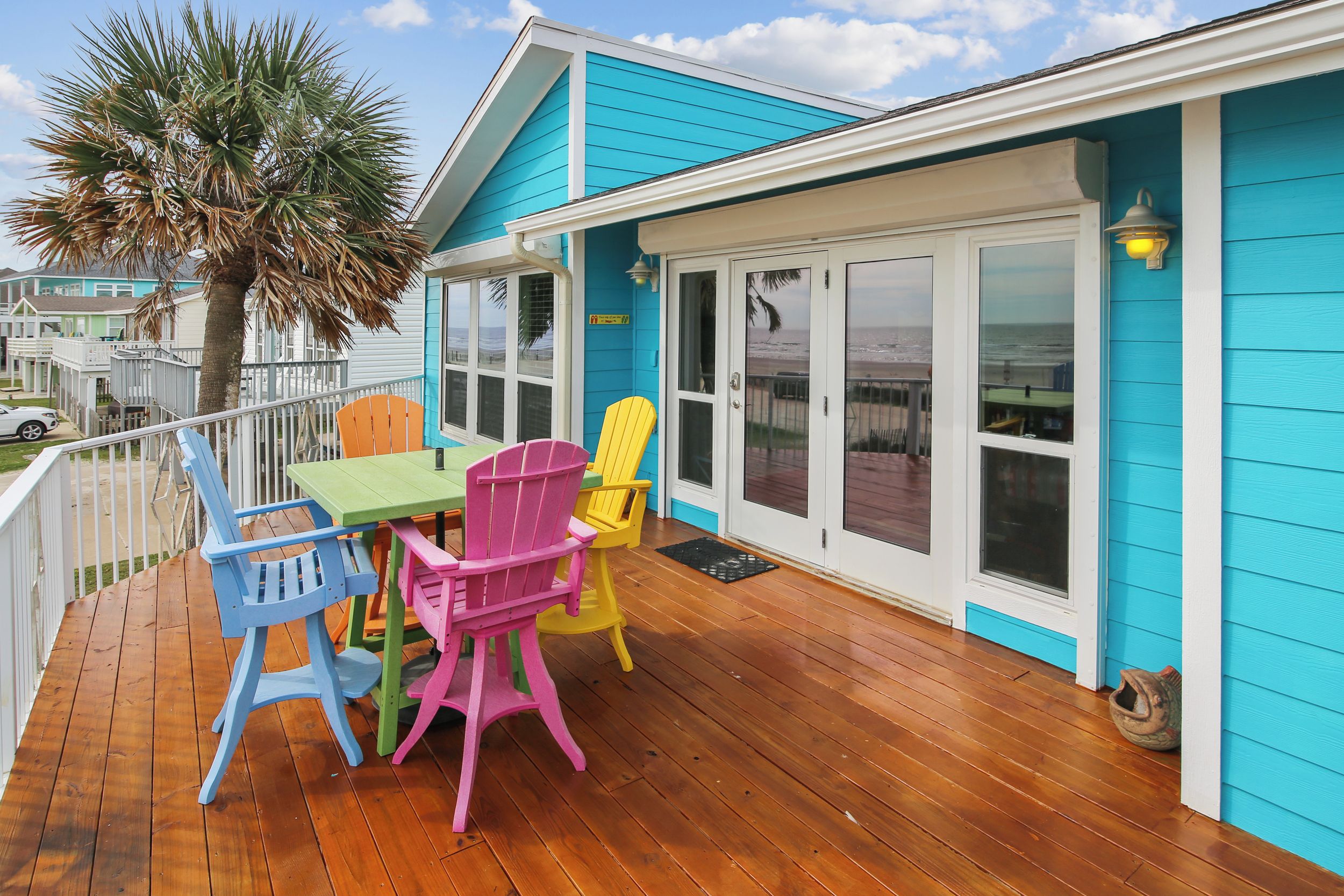 Um deck com mesa e cadeiras coloridas, uma palmeira e uma casa com revestimento azul e detalhes brancos.