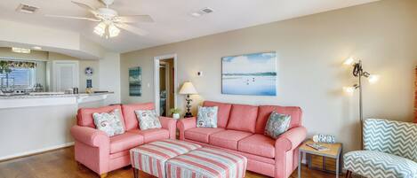 A living room with a pink sofa set, a striped ottoman, a small side table, a floor lamp, and a painting of a beach scene.