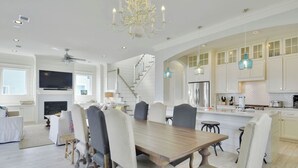 Un comedor amplio con una gran mesa de madera y sillas tapizadas, una cocina con armarios blancos y lámparas colgantes azules, y una escalera que sube a un nivel superior.