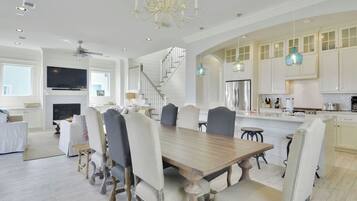 A spacious dining area with a large wooden table and upholstered chairs, a kitchen with white cabinets and blue pendant lights, and a staircase leading to an upper level.