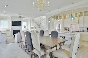 A spacious dining area with a large wooden table and upholstered chairs, a kitchen with white cabinets and blue pendant lights, and a staircase leading to an upper level. - Beachside Haven by Ryson Vacation Rentals (Galveston)