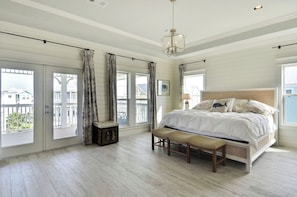 A bedroom with a large bed, a bench, a chest of drawers, and a view of the outside balcony. - Beachside Haven by Ryson Vacation Rentals (Galveston)