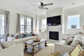 A spacious living room with a fireplace, a television mounted above it, and a large window with curtains. - Beachside Haven by Ryson Vacation Rentals (Galveston)
