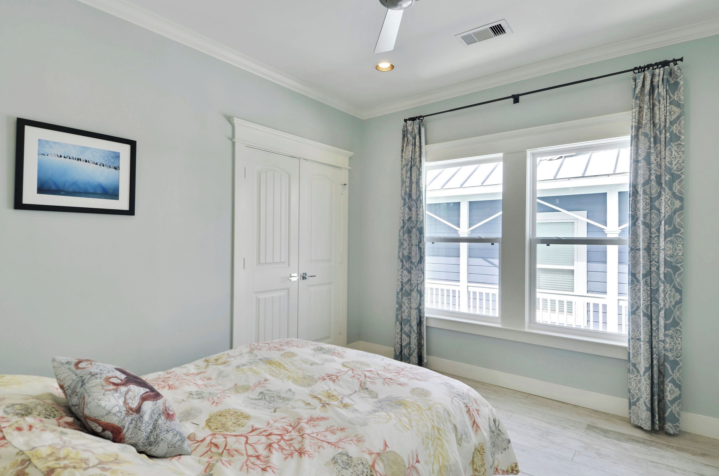 A bedroom with a bed, a framed picture on the wall, a white door, and large windows with curtains.