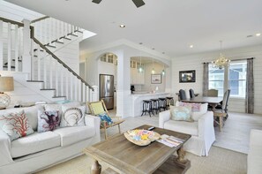 A spacious living room with a white staircase, a kitchen area, and a dining space with bar stools.