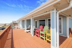 En träveranda med färgglada Adirondack-stolar, ett takförsett område med fönster och utsikt över stranden.
