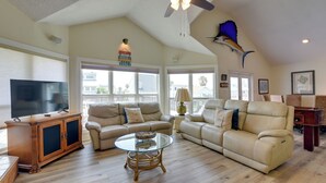 A spacious living room with a ceiling fan, a large flat-screen TV, two sofas, a glass coffee table, and a wooden cabinet. - 1 Mi to Sea Isle Beach: Canal-Front Gem w/ Dock! (Galveston)