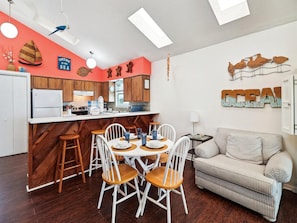 A modern kitchen with wooden cabinets, a white countertop, and a dining area with white chairs and a striped sofa. - Bermuda Bungalow by Ryson Vacation Rentals (Galveston)