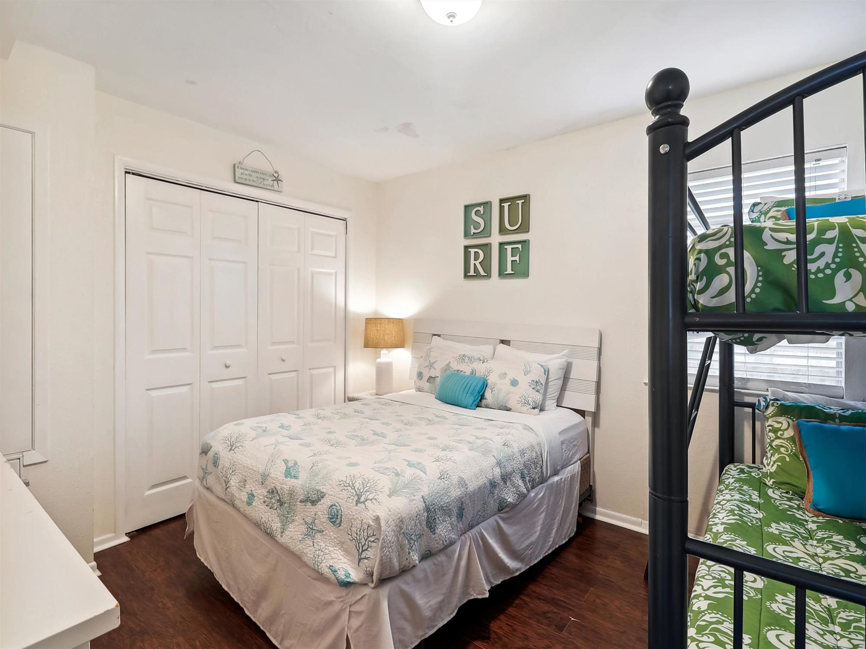 A bedroom with a bed, a nightstand, a lamp, a bunk bed with patterned bedding, a closet, and a wall with 'SURF' letters.