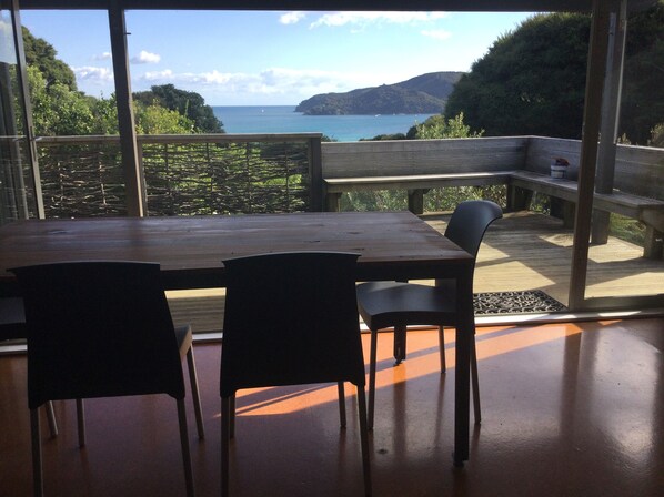 A wooden table with four chairs in a room with large windows overlooking a coastal landscape. - Cottage,2 Bedroom,Tryphena,Great Barrier Island.Great Location (Auckland)