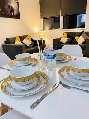 A dining table set with white plates and gold accents, surrounded by modern furniture and a large window.