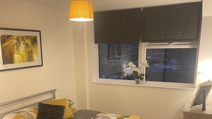 A neatly arranged bedroom with a bed, bedside table, and a window with a view of the city at night. - Modern city centre two bedroom fully equipped apartment (Derby)